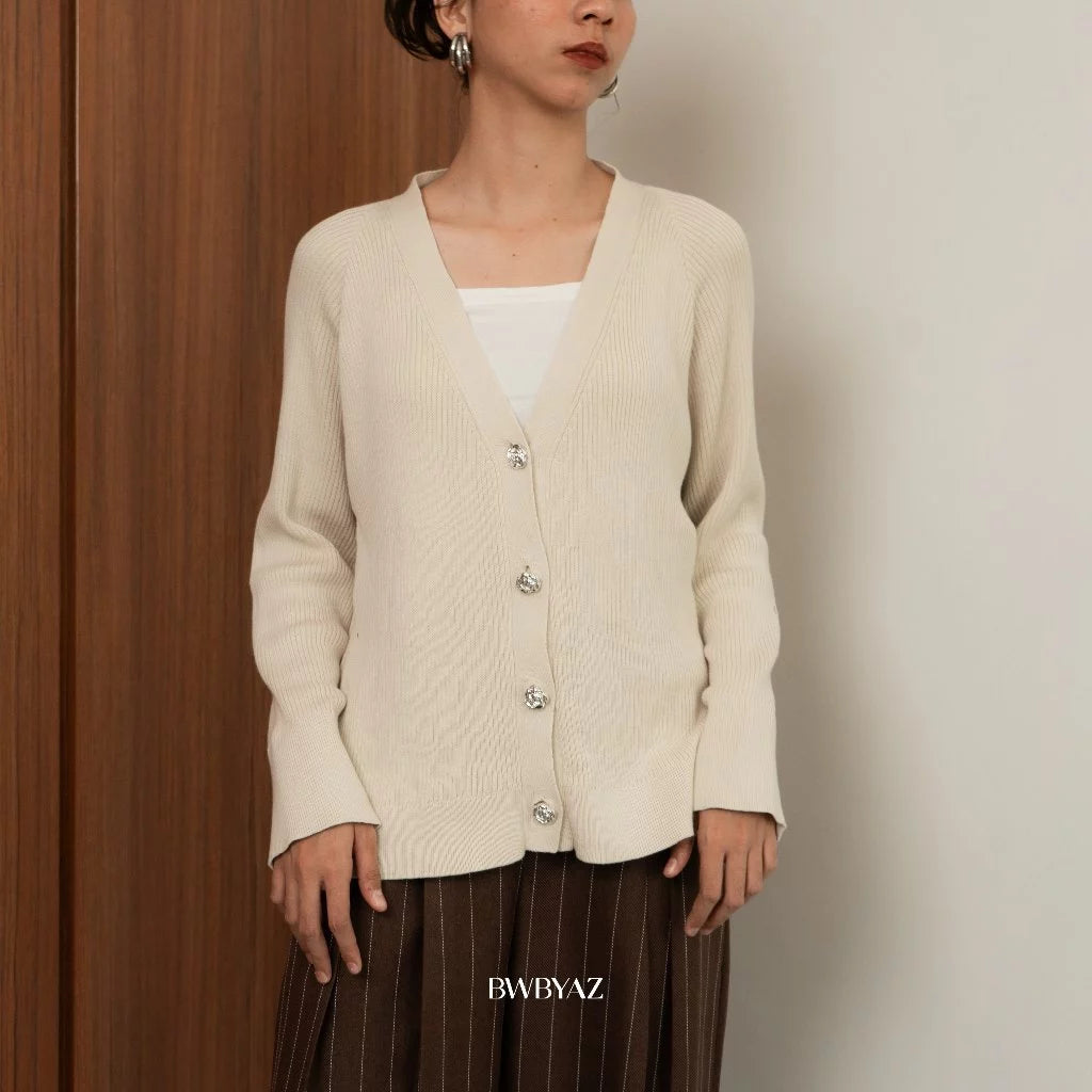 A woman wearing a beige knit cardigan with long sleeves and silver buttons from BWBYAZ, paired with a white top and a brown skirt.