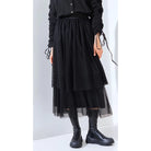 A person wearing a layered black skirt by Yestoday with a elastic waistband and black boots. The skirt has a solid color and appears to be made of a smooth fabric with a flared design.