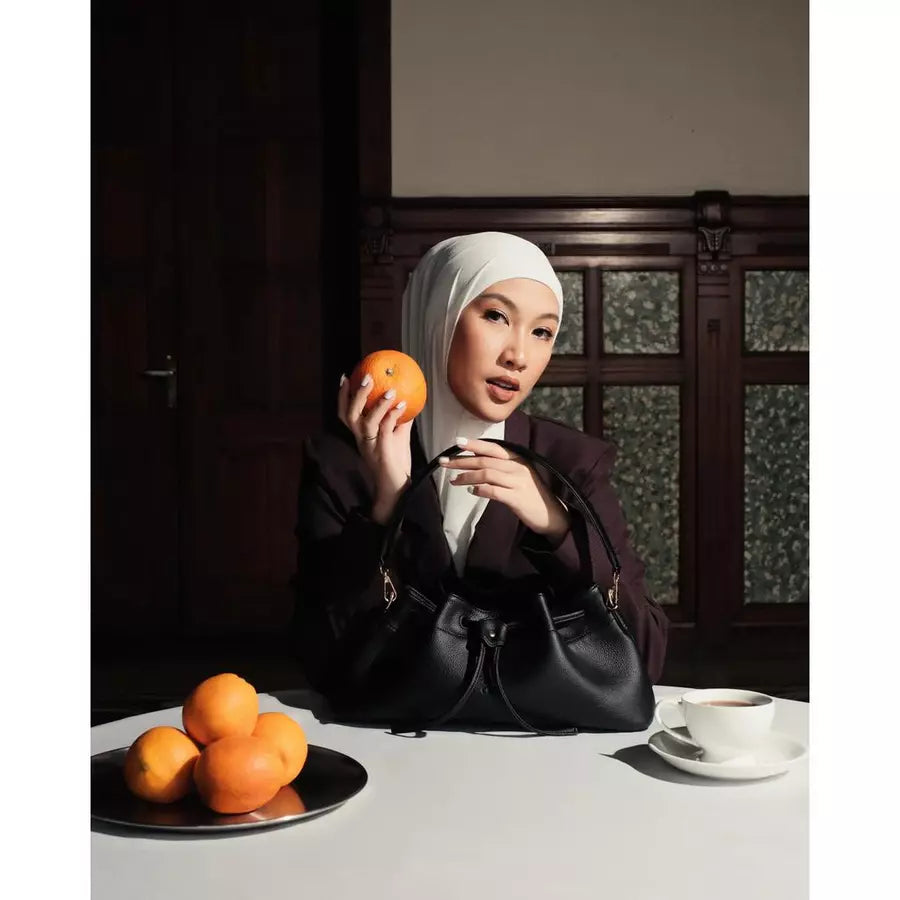 Woman holding black shoulder bag by Prior at a table with oranges and a cup of coffee.