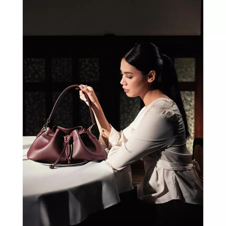 Woman sitting at a table with a burgundy shoulder bag by Prior in a dimly lit room