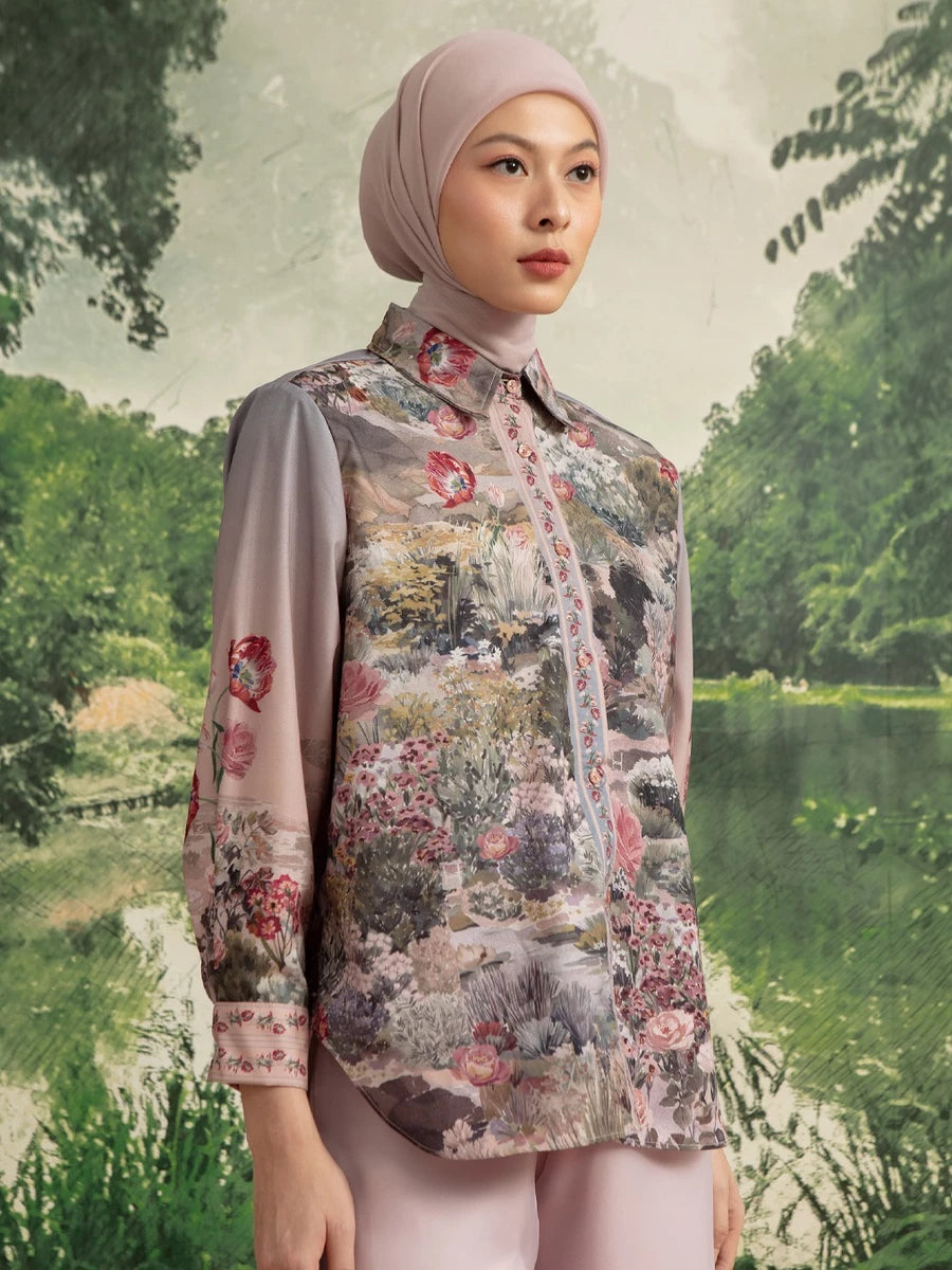 A woman wearing a light pink hijab and a matching long-sleeve button-up shirt by Hijabchic with a detailed multicolor landscape print on the front and sleeves, set against a scenic forest background.