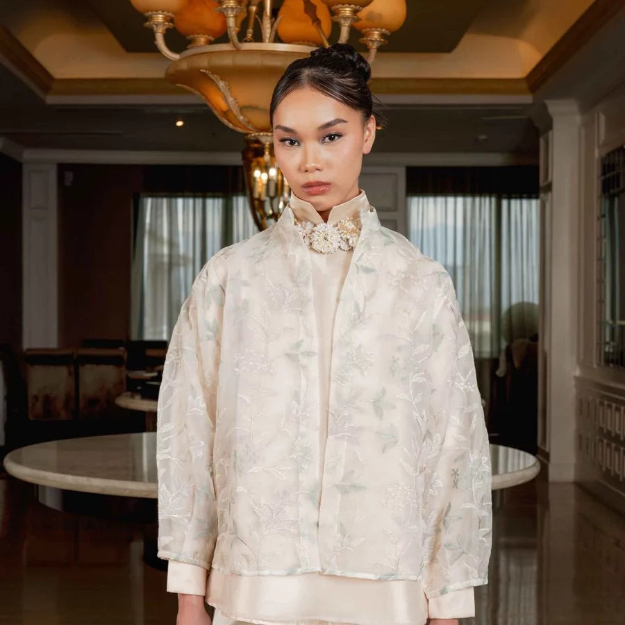 A woman stands indoors wearing an ash grey-colored embroidered jacket by Jenna & Kaia with a high collar and a decorative floral applique at the neck, paired with a matching cream satin garment. The setting is a luxurious interior with warm lighting, chandeliers, and elegant furniture.