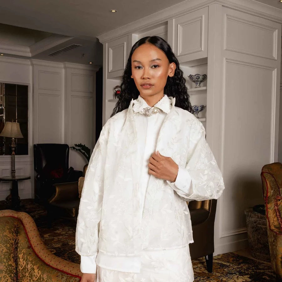 A woman stands indoors wearing a broken white-colored embroidered jacket by Jenna & Kaia with a high collar and a decorative floral applique at the neck, paired with a matching cream satin garment. The setting is a luxurious interior with warm lighting, chandeliers, and elegant furniture.