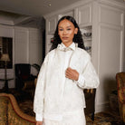 A woman stands indoors wearing a broken white-colored embroidered jacket by Jenna & Kaia with a high collar and a decorative floral applique at the neck, paired with a matching cream satin garment. The setting is a luxurious interior with warm lighting, chandeliers, and elegant furniture.