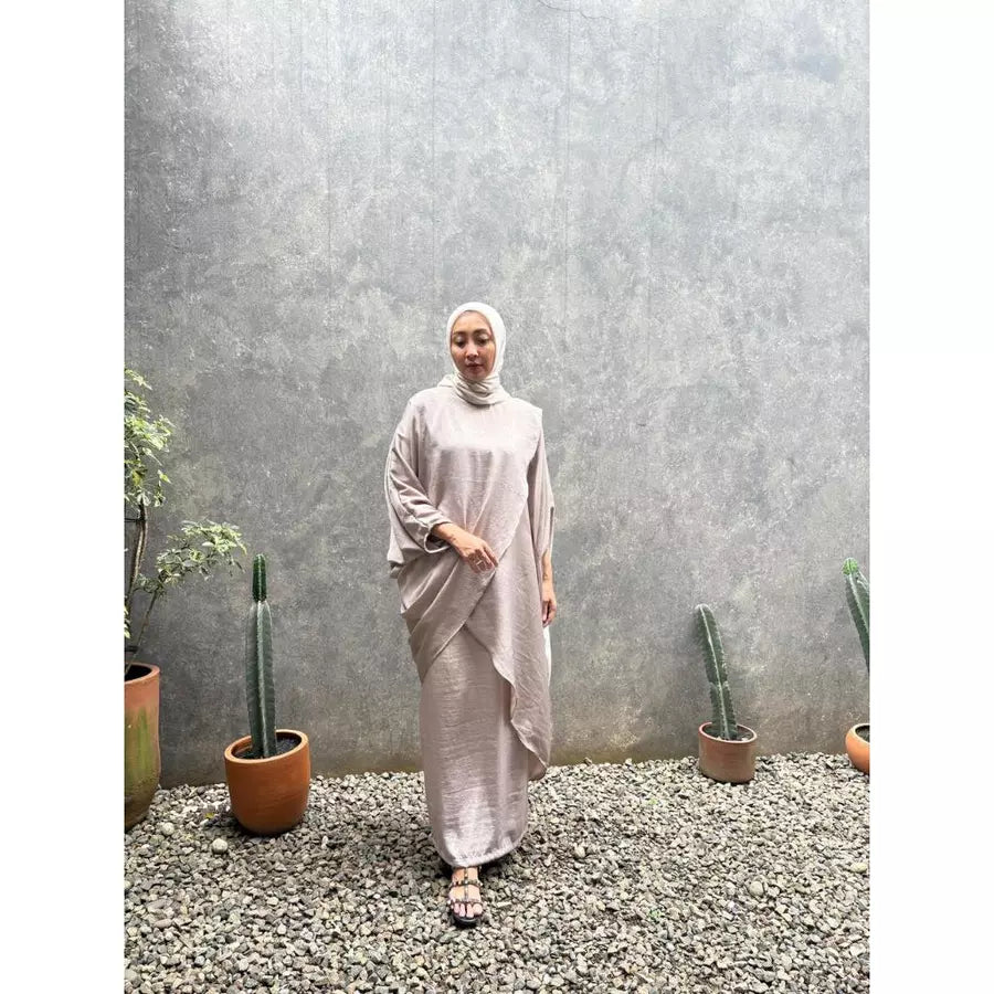 Woman in a light-colored outfit set by Abame standing in front of a textured wall with potted plants.