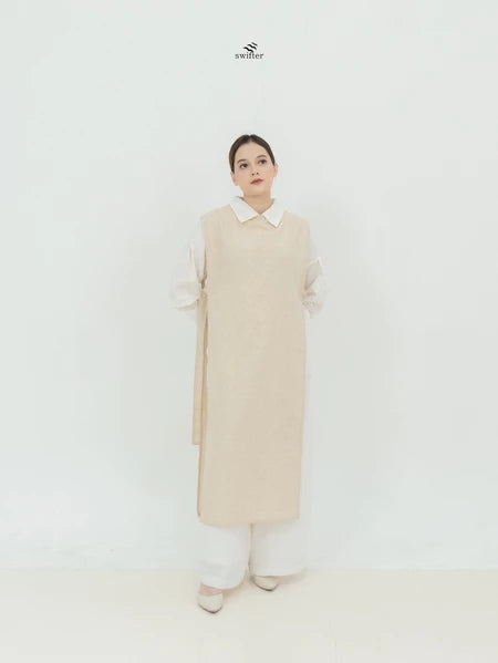 A person standing in a studio setting, wearing a long beige vest by Swifter over a white shirt and white pants.