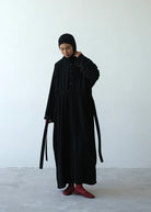 A person wearing a black traditional abaya by Rashawl with a drawstring hood standing against a plain background.