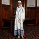 Woman in a long, white floral dress with light blue accents by Lozy Hijab standing in a room with wooden paneling.