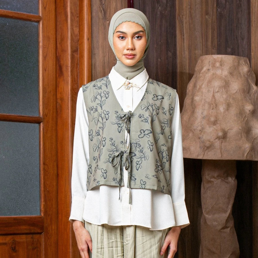 Woman wearing a patterned sage vest by Lozy Hijab over a white shirt in front of a wooden door.