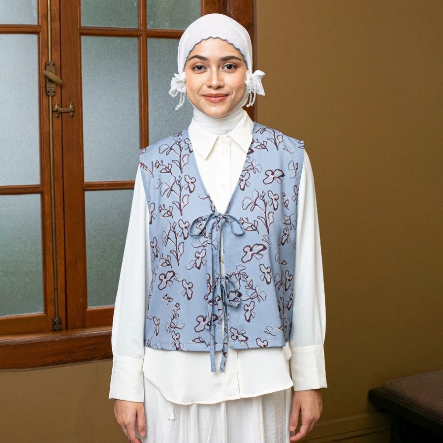 Woman wearing a floral sky blue vest by Lozy Hijab over a white shirt in a room with a wooden door.
