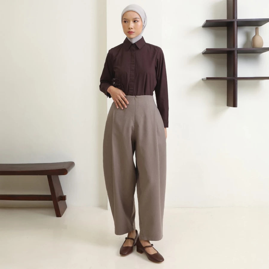 Woman wearing a brown shirt and mocca wide-leg pants by Hijabchic standing in a minimalistic room.