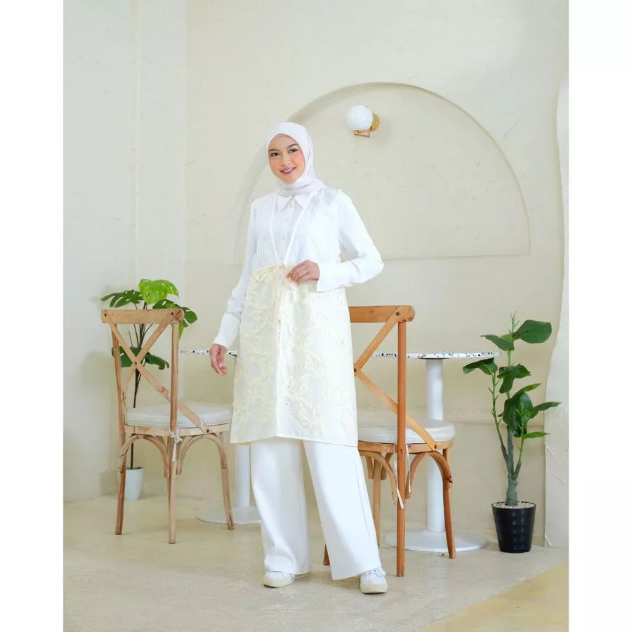 Woman wearing a white vest tunic by Abame standing in a minimalistic room with plants and chairs.