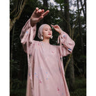 A woman wearing a long, printed dress by Maima with long sleeves in a dusty color, standing on a pathway in a forested area.