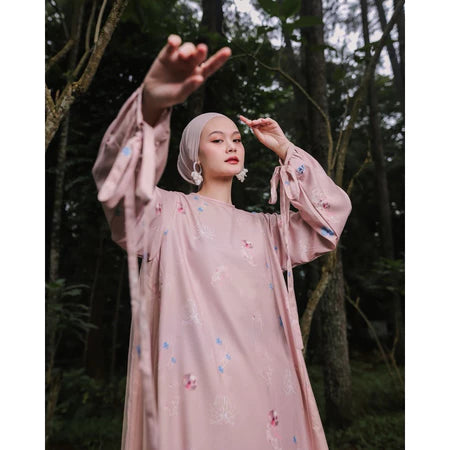 A woman wearing a long, printed dress by Maima with long sleeves in a dusty color, standing on a pathway in a forested area.