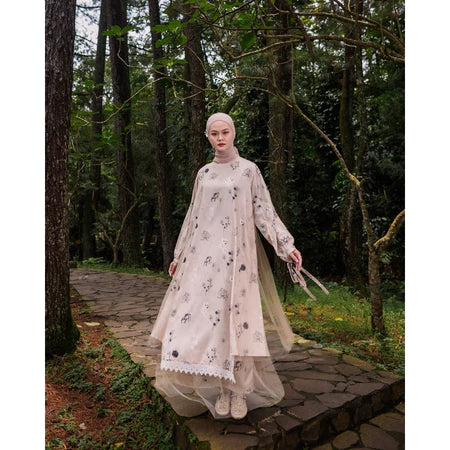 A woman wearing a long, printed dress by Maima with long sleeves in a mocca color, standing on a pathway in a forested area.