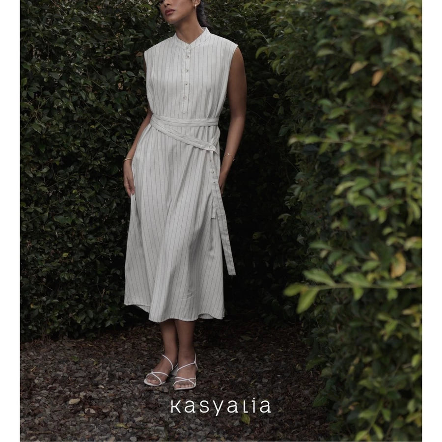Woman wearing a light-colored dress with a belt standing in a garden setting, with 'kasyalia' branding.