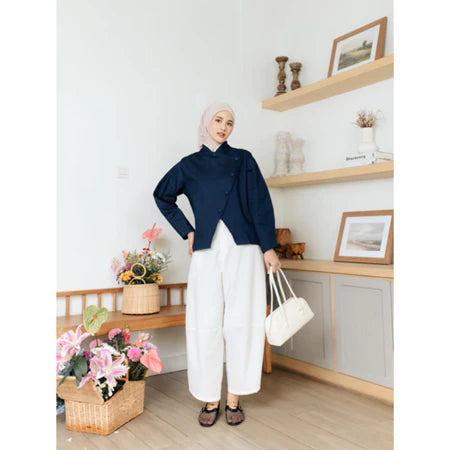 Woman in navy top from Haidee Orlin and white pants holding a white bag in a room with wooden shelves and flowers.