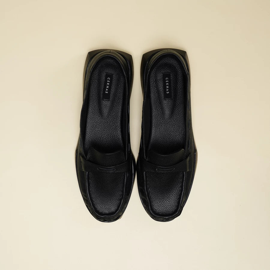 A pair of black leather penny loafers flatshoes with a chunky black sole, photographed from a top-down view on a light beige background. The brand name 'Cekhas' is visible on the insole.