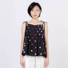A model wearing a black sleeveless top by Kasyalia with white polka dots and white pants, standing against a plain light gray background.