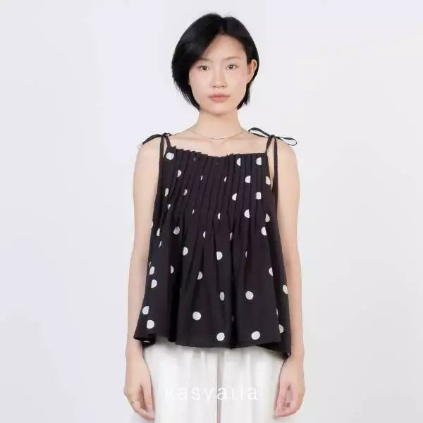 A model wearing a black sleeveless top by Kasyalia with white polka dots and white pants, standing against a plain light gray background.