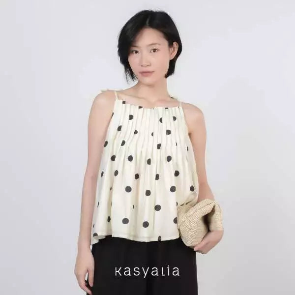 A woman modeling a cream-colored sleeveless top with black polka dots, paired with black pants and holding a woven straw clutch bag. The brand name 'kasyalia' is printed at the bottom.