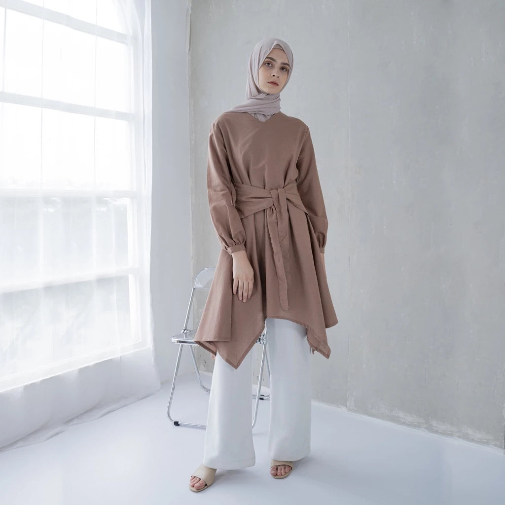 A woman wearing a brown tunic with a V-neck and pleats on the sleeves, paired with white pants, standing in a bright room.