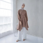 A woman wearing a brown tunic with a V-neck and pleats on the sleeves, paired with white pants, standing in a bright room.