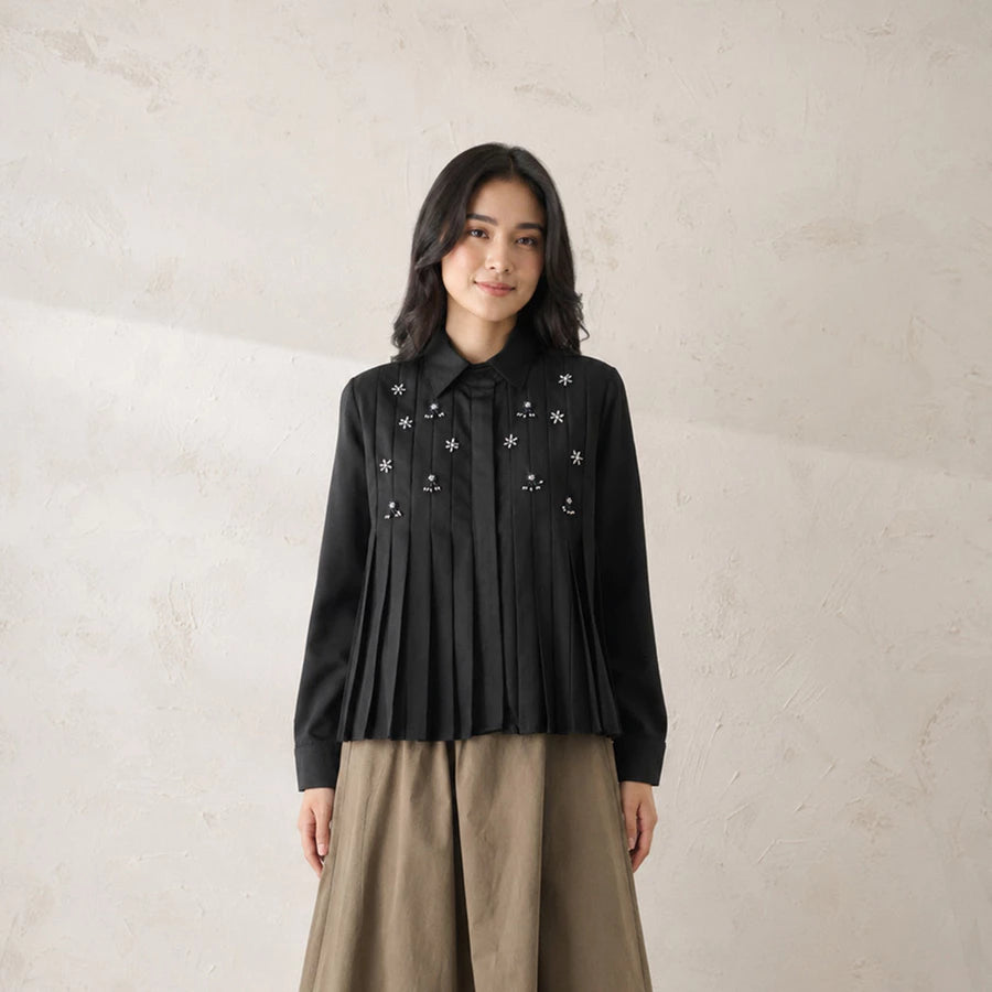 A woman stands against a textured beige wall wearing a black long-sleeve pleated shirt by Jenna & Kaia with embroidered white floral details and a bow at the collar, paired with a beige skirt.