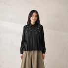 A woman stands against a textured beige wall wearing a black long-sleeve pleated shirt by Jenna & Kaia with embroidered white floral details and a bow at the collar, paired with a beige skirt.