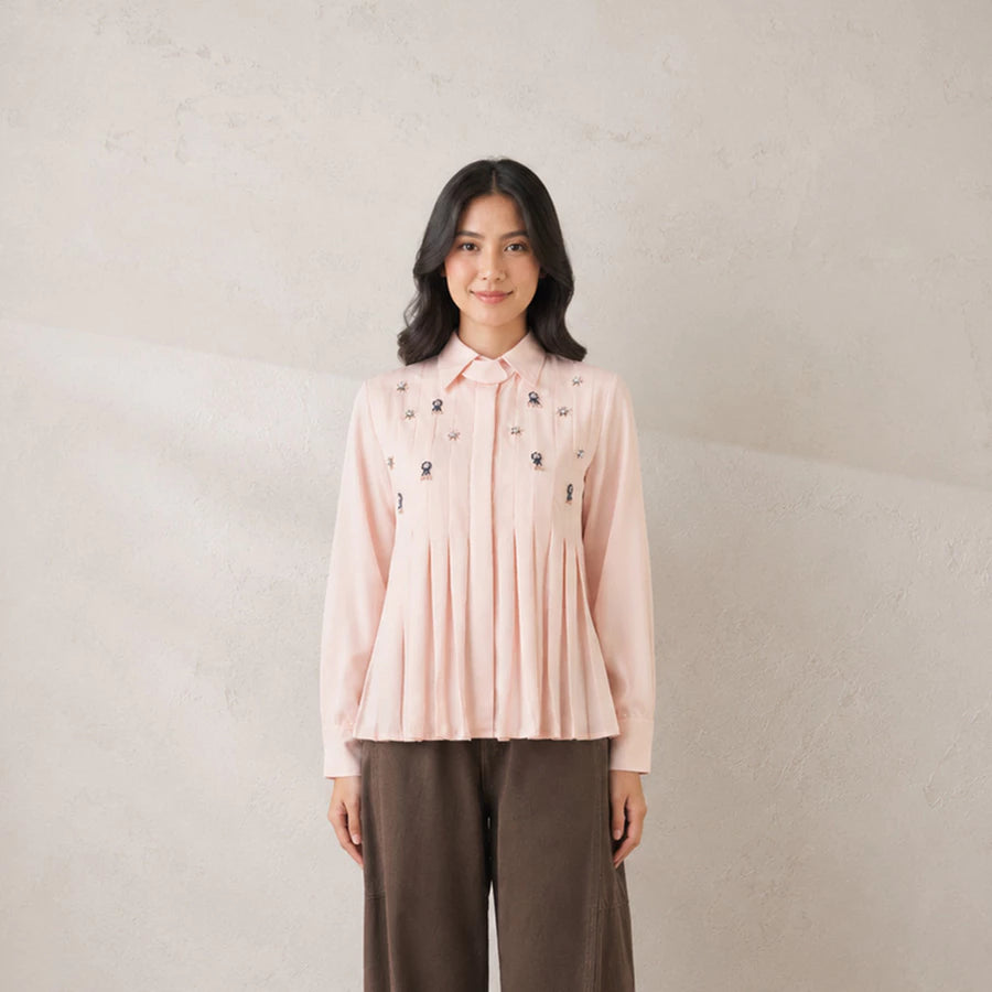 A woman stands against a textured light beige wall wearing a dusty pink pleated shirt by Jenna & Kaia with small dark embroidered details on the chest and dark brown wide-leg pants.