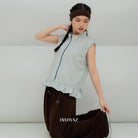 A model wearing a light aqua blue sleeveless vest by BWBYAZ with a ruffled hem and dark brown pants, kneeling against a minimalist light gray background. The brand name 'BWBYAZ' is printed on the pants.