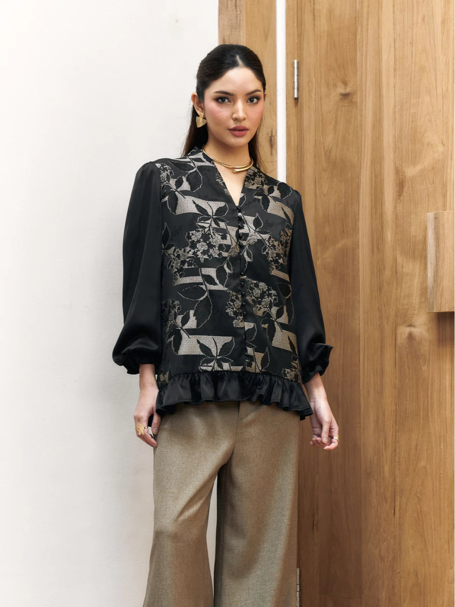 Woman wearing a black floral blouse by Studio Tui and beige pants against a wooden door.