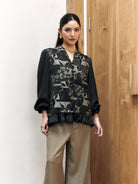 Woman wearing a black floral blouse by Studio Tui and beige pants against a wooden door.