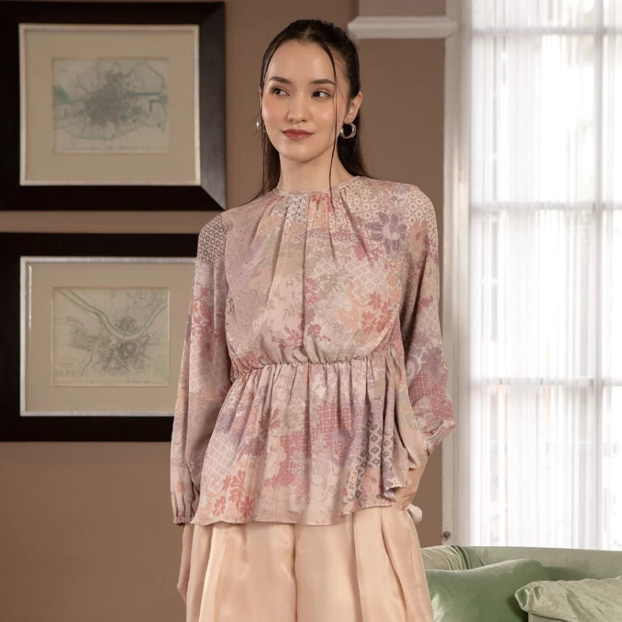 Woman wearing a peach-colored floral blouse by Hijabchic standing in a room with framed artwork on the wall.