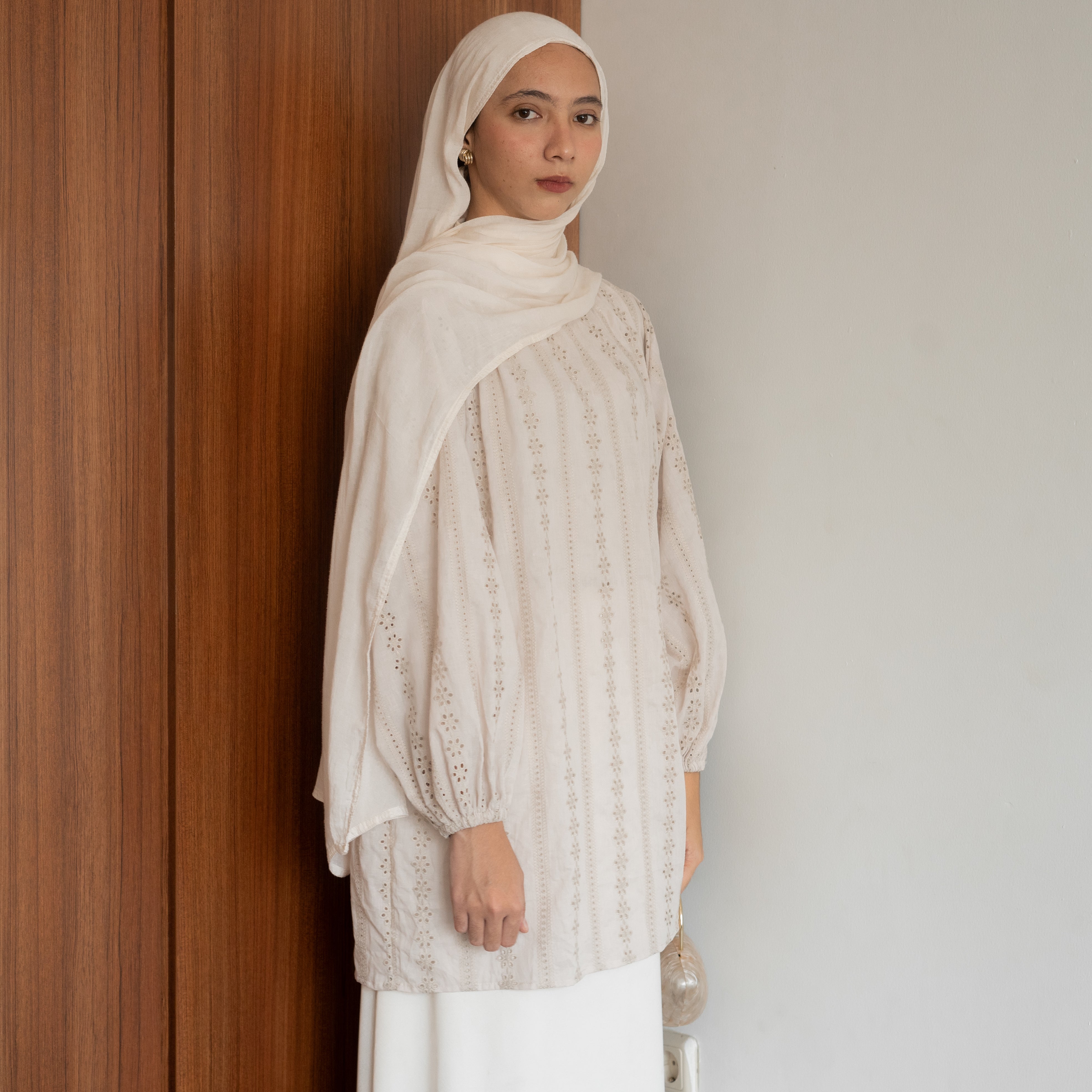 A woman wearing an elegant premium cotton lace tunic in cream from BWBYAZ, styled with a matching hijab and ivory skirt. The reversible tunic features delicate lace details, tie accents at the neckline, and bishop sleeves with elastic cuffs, paired with a small metallic gold handbag for a refined, versatile, and modest fashion look.