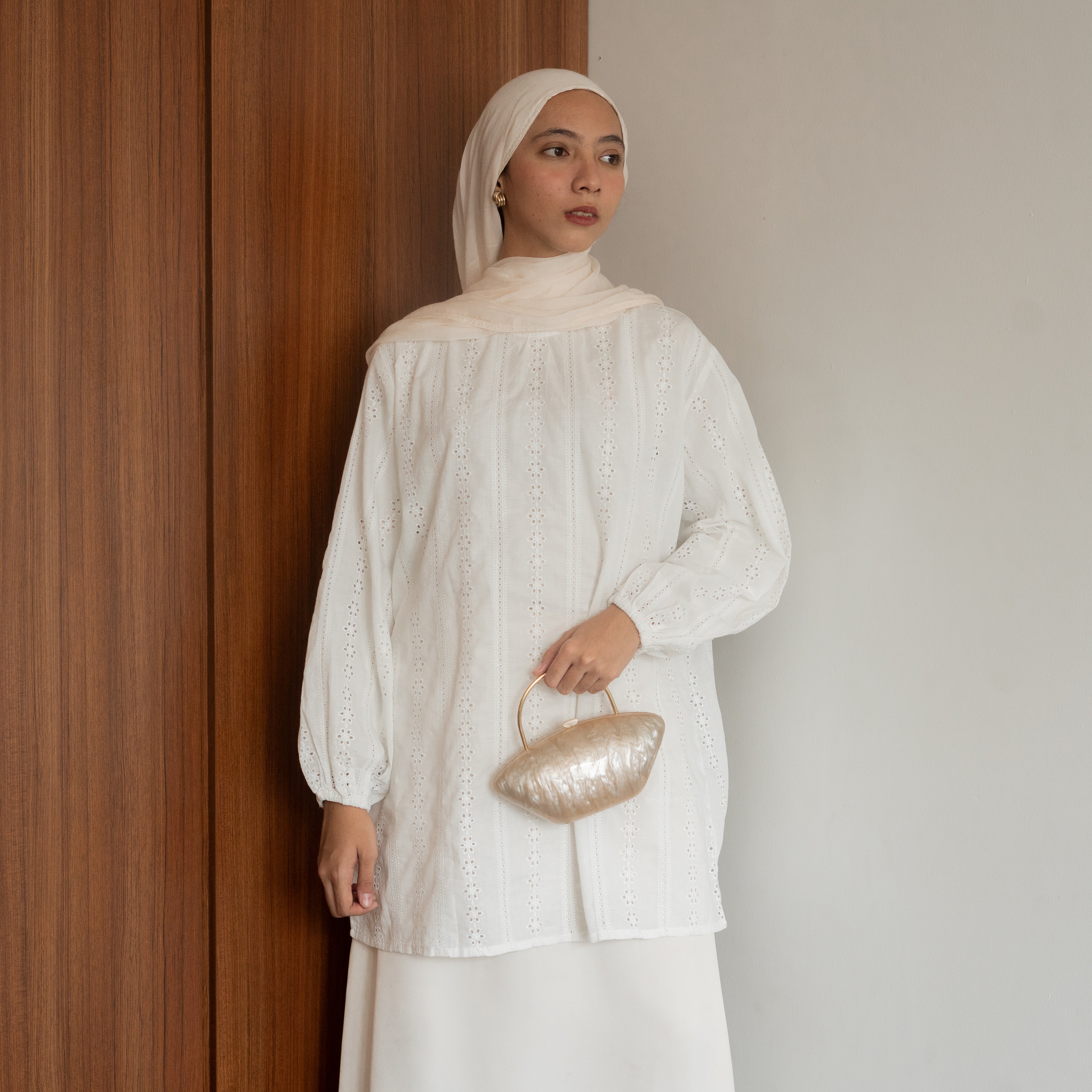 A woman wearing an elegant premium cotton lace tunic in white from BWBYAZ, styled with a matching hijab and ivory skirt. The reversible tunic features delicate lace details, tie accents at the neckline, and bishop sleeves with elastic cuffs, paired with a small metallic gold handbag for a refined, versatile, and modest fashion look.