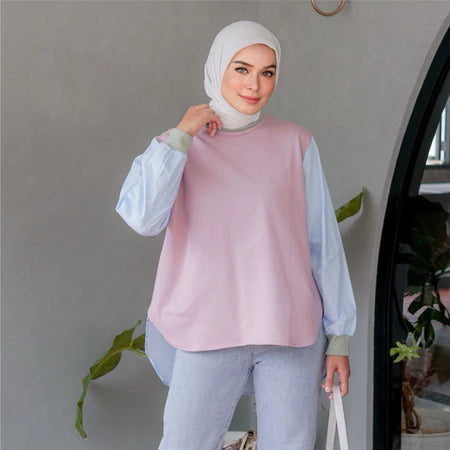 Woman wearing a light pink shirt with light blue sleeves by Casa Elana and light blue pants, standing indoors.
