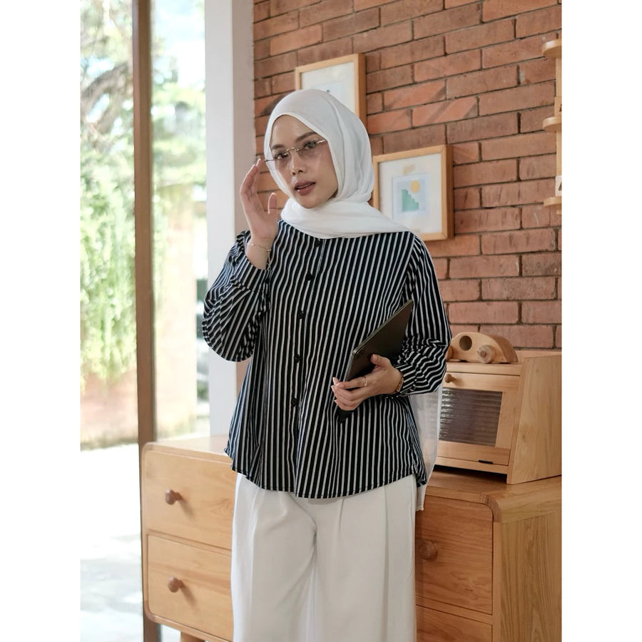 Woman wearing a black and white striped shirt by Renaette and hijab standing in a room with wooden furniture and a brick wall.