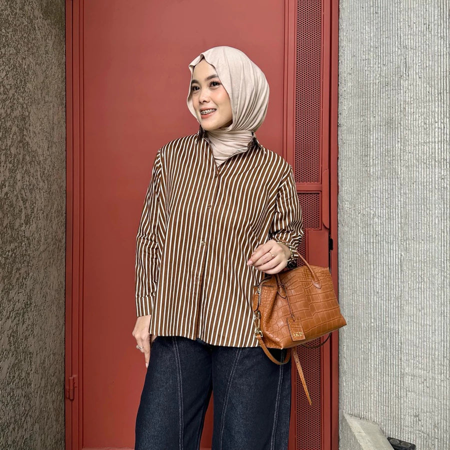 Woman wearing a brown striped shirt by Renaette and hijab, holding a brown leather bag against a red door.