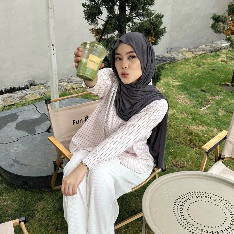 Woman in pink striped shirt by Renaette sitting outdoors holding a green cup, with a casual setting in the background.