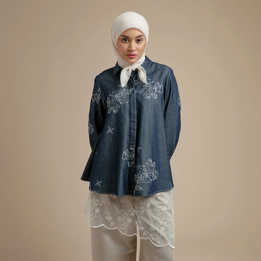 Woman wearing a dark denim shirt by Hijabchic with floral embroidery and a white hijab on a beige background