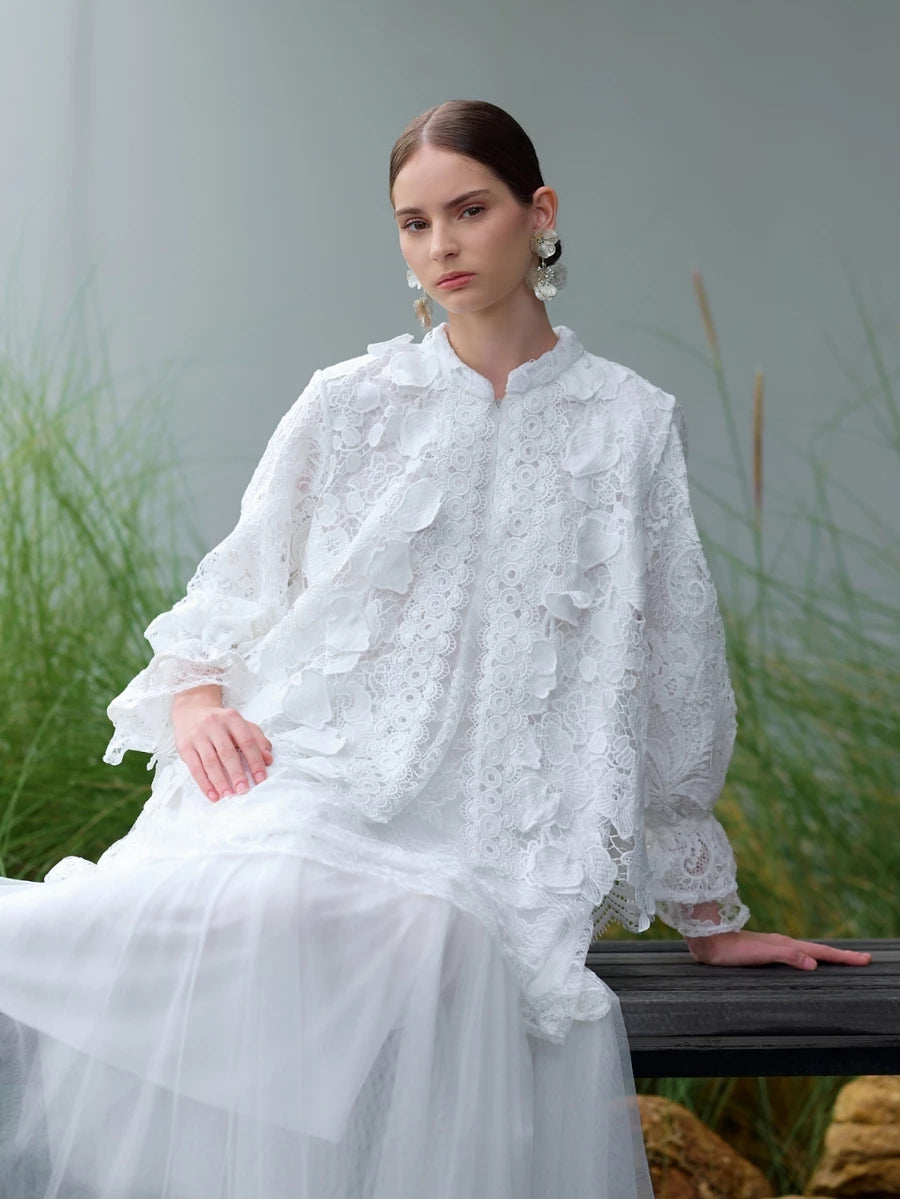 Woman in a white lace blouse by Kasa Heritage sitting outdoors with greenery in the background.