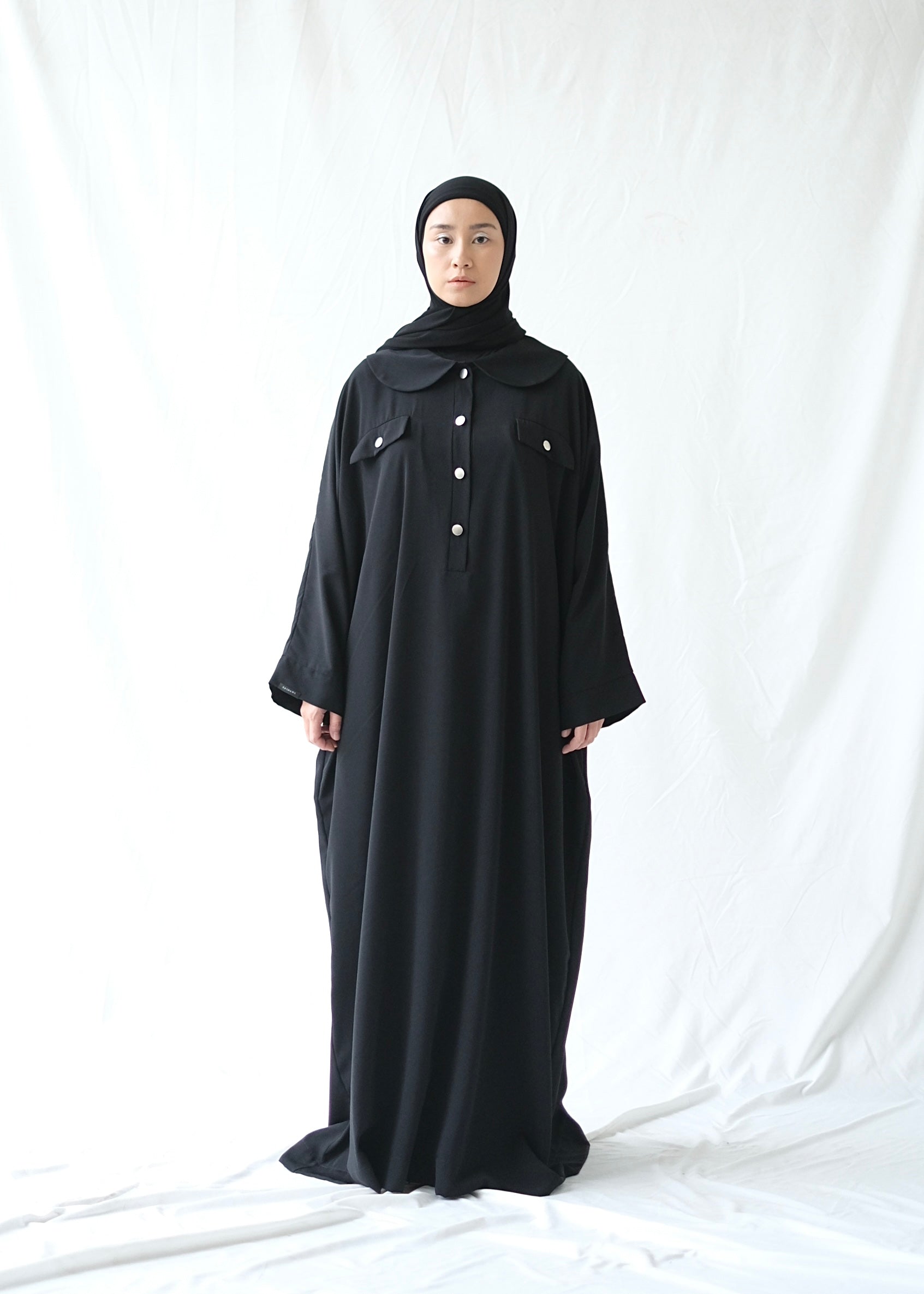 A person wearing a black abaya by Rashawl with a button opening on the front, standing against a white background.