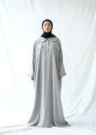 A person wearing a grey abaya by Rashawl with a button opening on the front, standing against a white background.