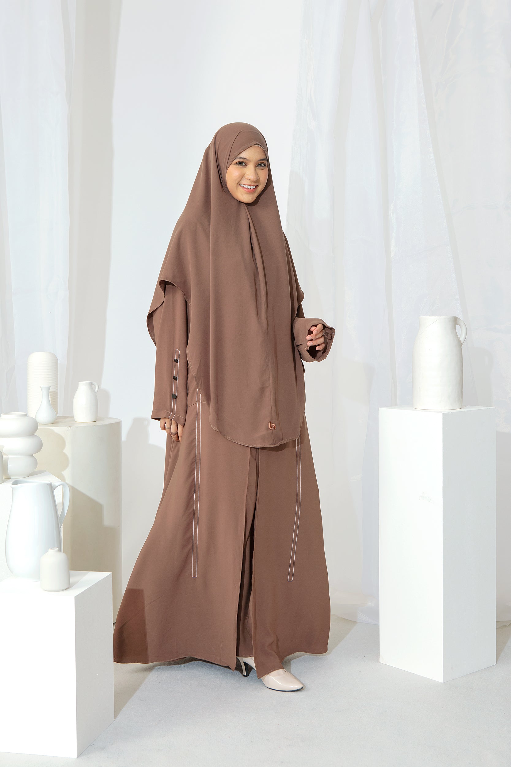 A woman wearing a brown Lozy Hijab abaya, standing against a neutral background with decorative candles to the side.