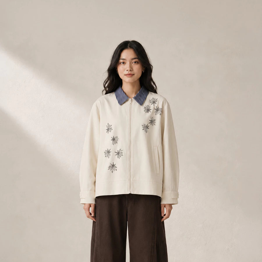 A woman stands against a warm beige wall wearing a cream-colored zip-up jacket by Jenna & Kaia with gray embroidered starburst patterns, a blue-and-white striped collar, and dark brown wide-leg pants.