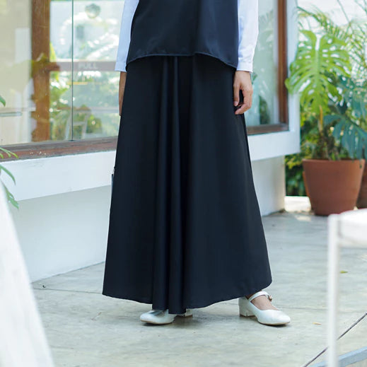 Person wearing a long black skirt by Abame with white shoes indoors.