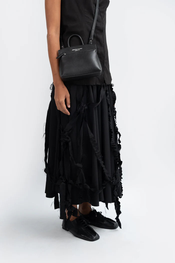 Person wearing a black ruffled skirt and black shoes with a black grain leather handbag by Aesthetic Pleasure on a white background