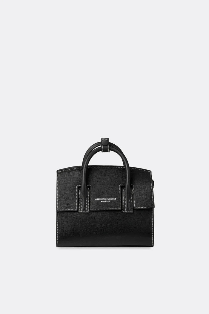 A mini-sized black grain leather handbag with a detachable strap and silver-tone hardware, featuring a front logo stamp and a tubular handle by Aesthetic Pleasure.
