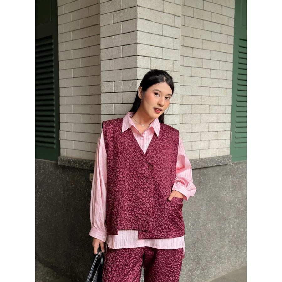 Woman wearing a maroon patterned vest over a pink shirt and pants, standing against a brick wall.